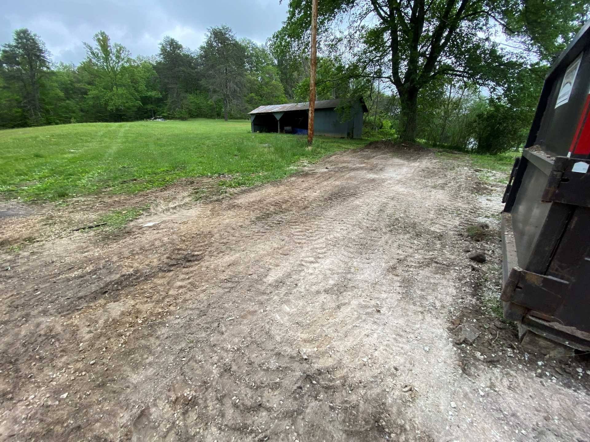 A gravel path leads toward a small shed in a grassy field under an overcast sky, with a dumpster in the foreground.