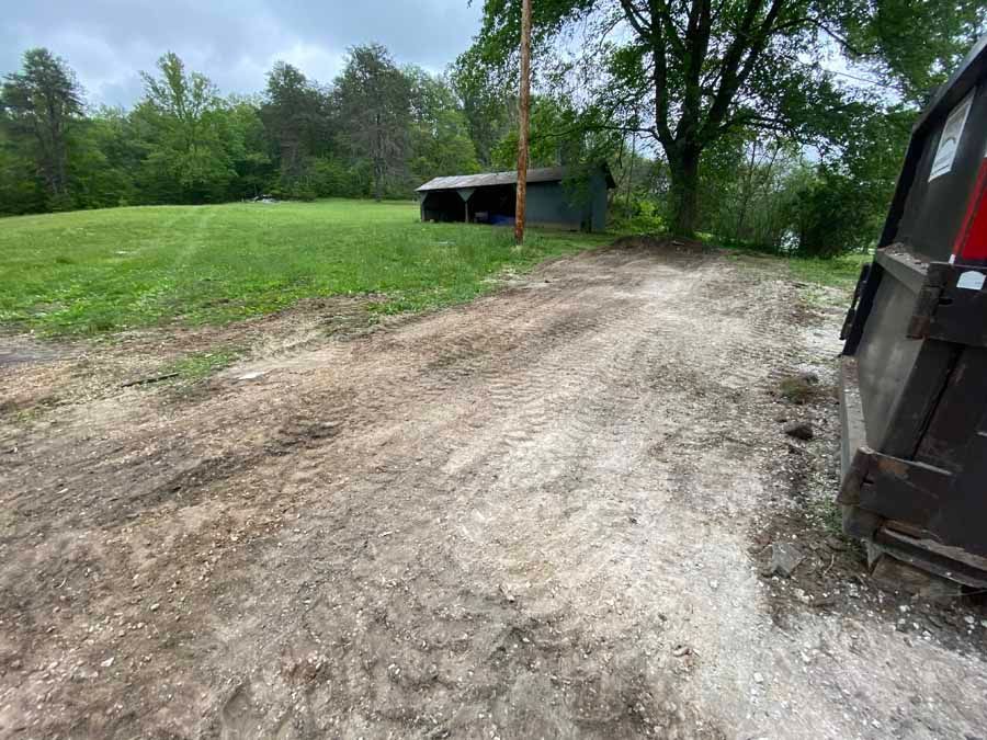 A gravel driveway leading toward a small shed in a grassy field under a cloudy sky, with a dumpster on the right.