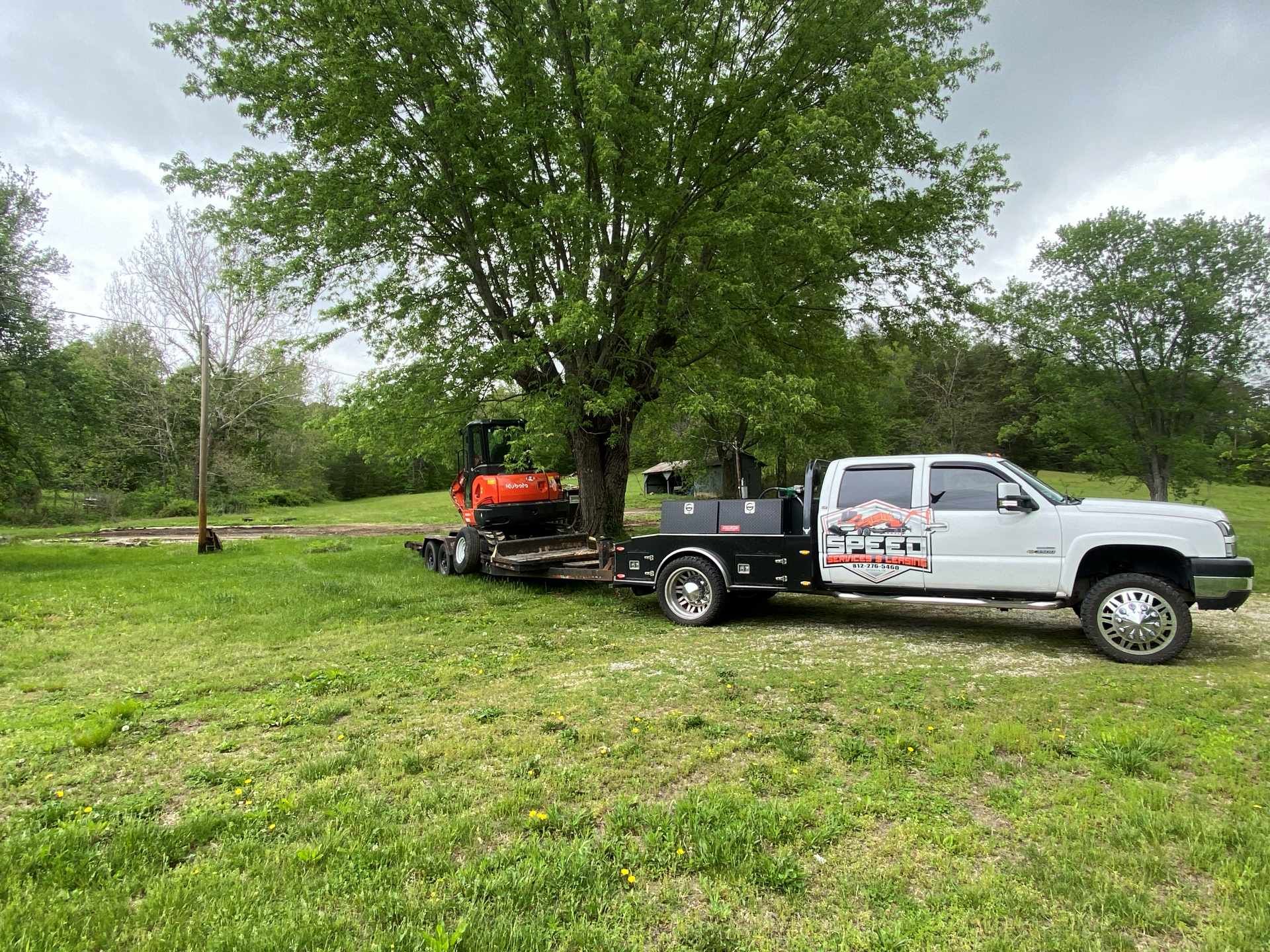 A white utility truck towing a trailer with an orange mini excavator parked under a large tree in a grassy field.