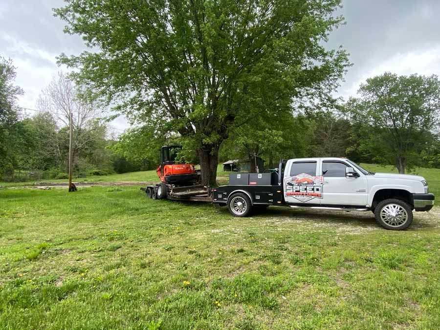 A white flatbed truck towing an orange excavator across a grassy field near a large tree.