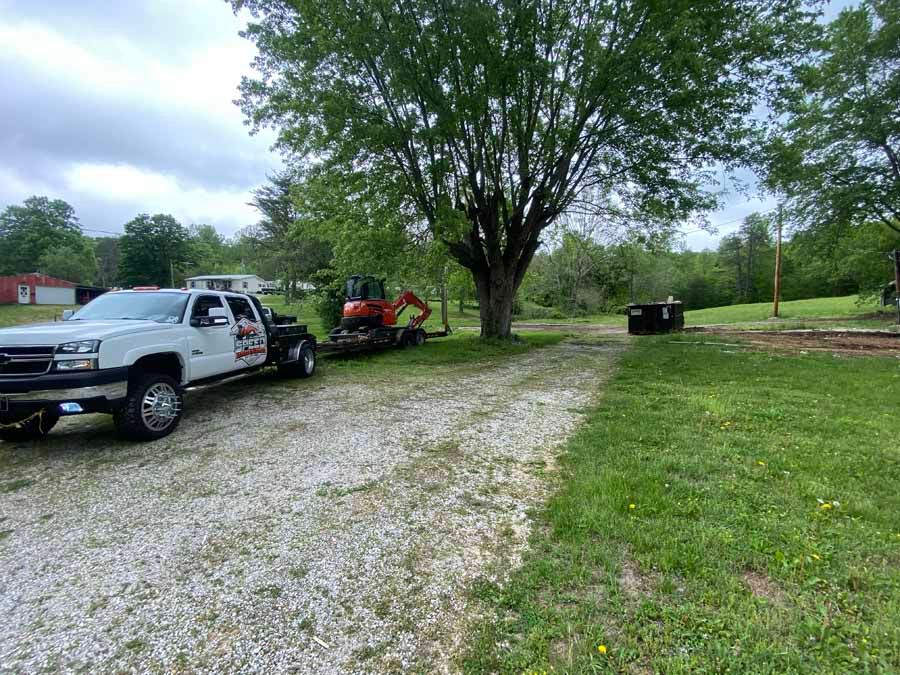A white pickup truck pulling a trailer with an orange excavator parked on a gravel driveway near a large tree.