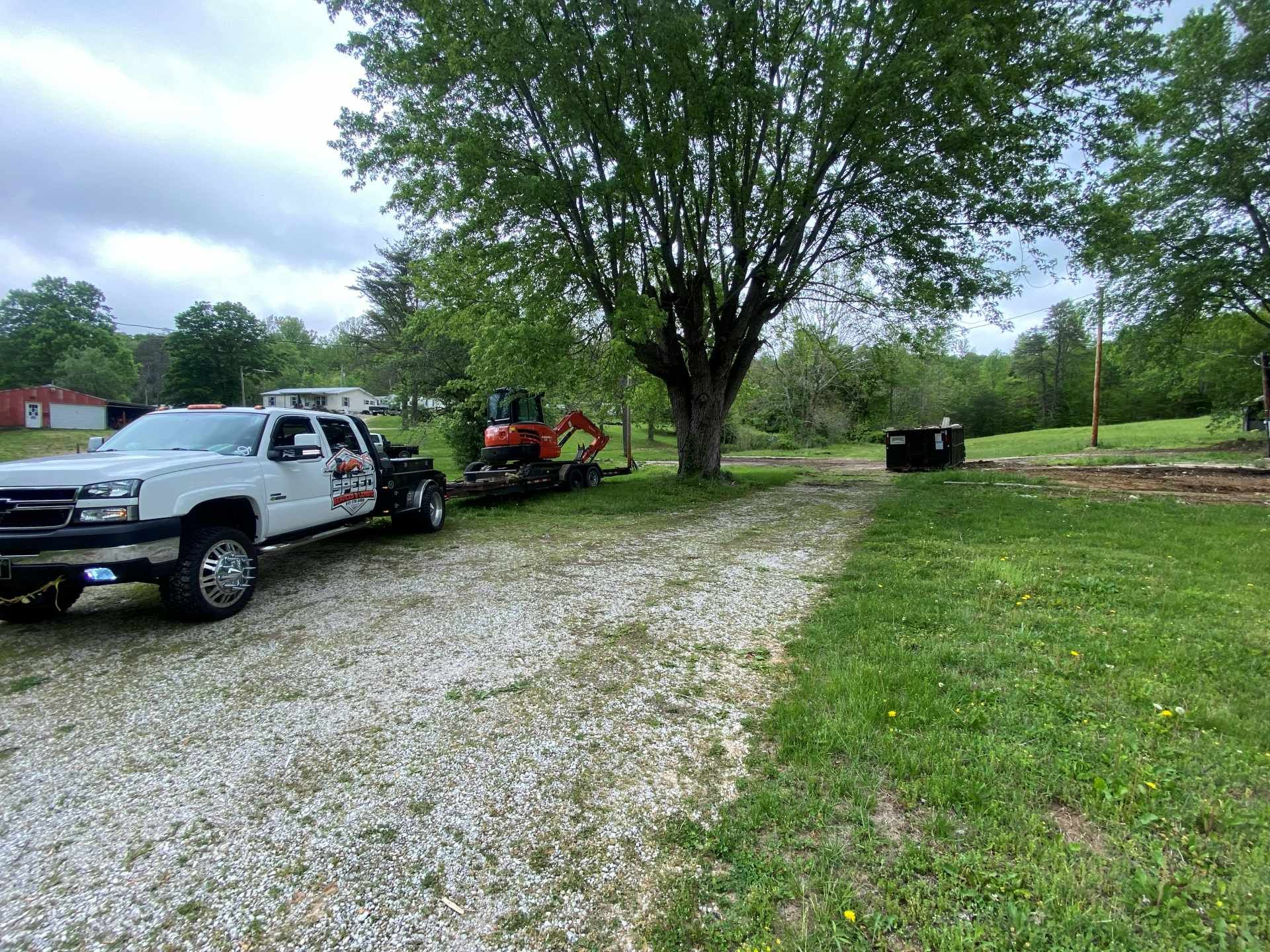 A white pickup truck pulling a trailer with a red excavator parked on a gravel driveway next to a large tree.