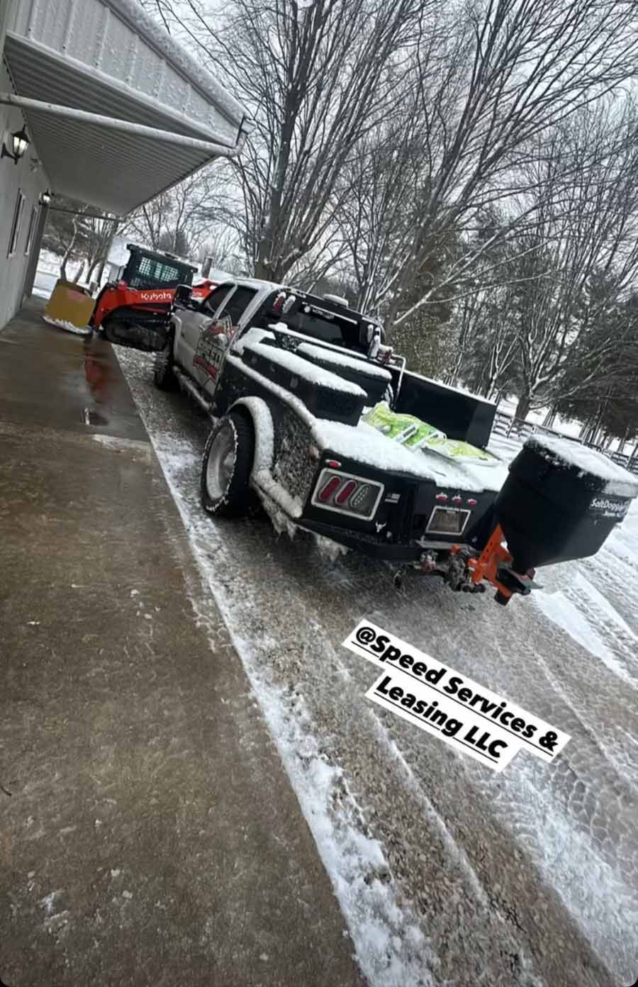 A snow-dusted work truck with a rear spreader attachment and a nearby skid steer parked by a building in winter.