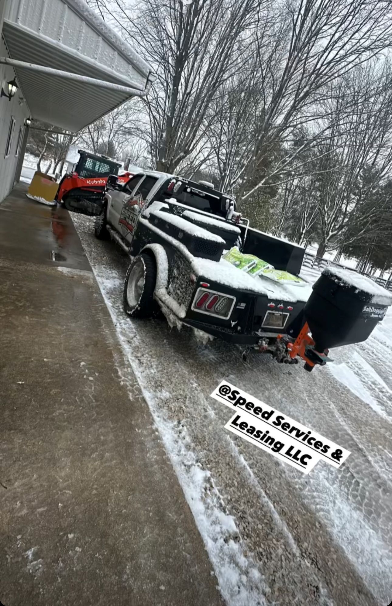 A pickup truck with a salt spreader attachment parked on a snowy lot next to a building, with a Bobcat loader nearby.