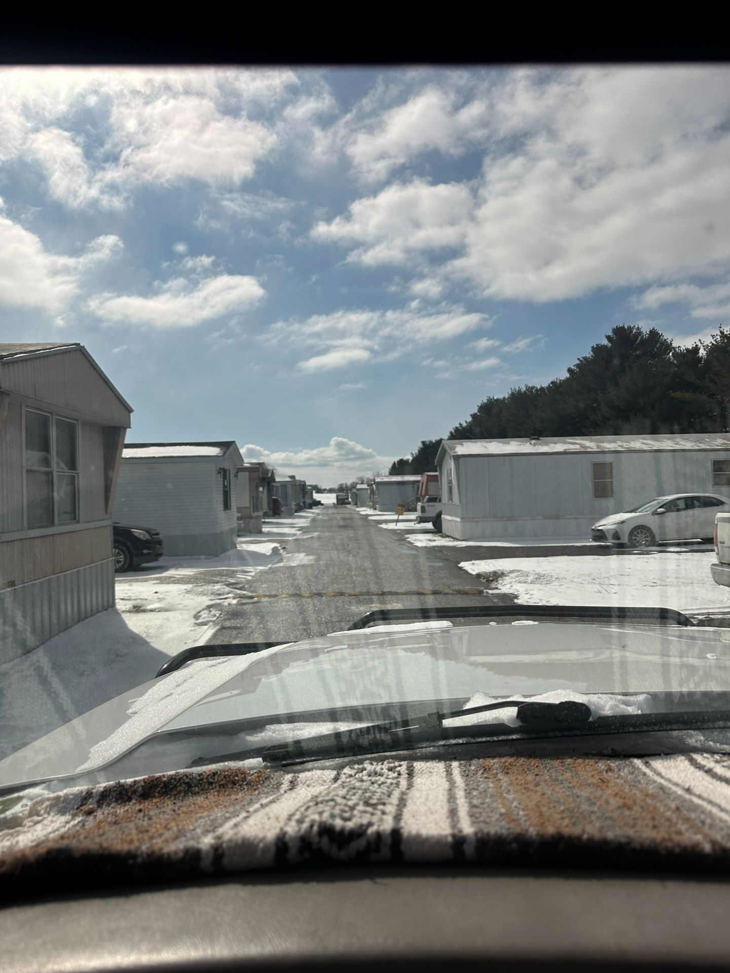 A view from a car windshield looking down a narrow street in a mobile home park covered in patchy snow on a sunny day.