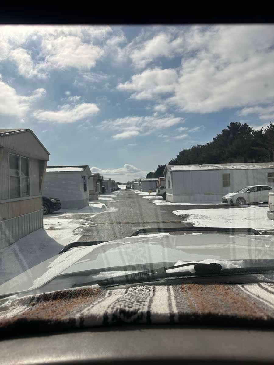 A view from inside a vehicle looking down a street lined with white mobile homes on a sunny, snow-covered day.