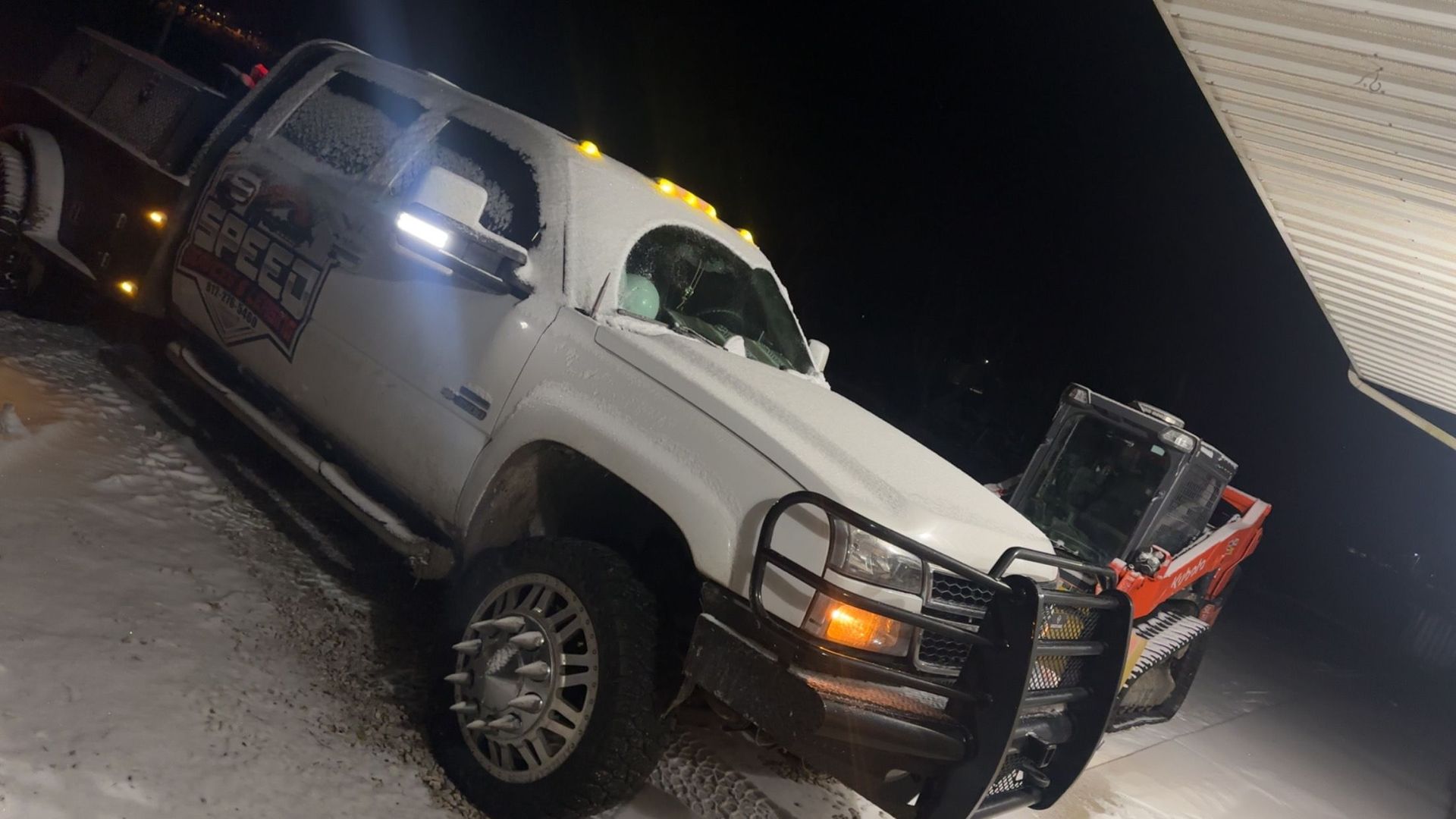 A white pickup truck with a brush guard parked in the snow at night, with a piece of orange machinery in the background.