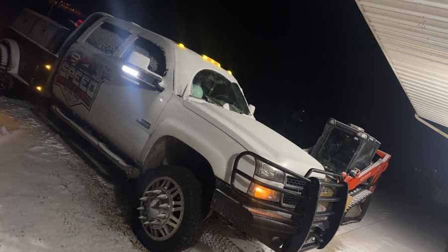 A white pickup truck with a brush guard parked in the snow at night, with a piece of orange machinery nearby.