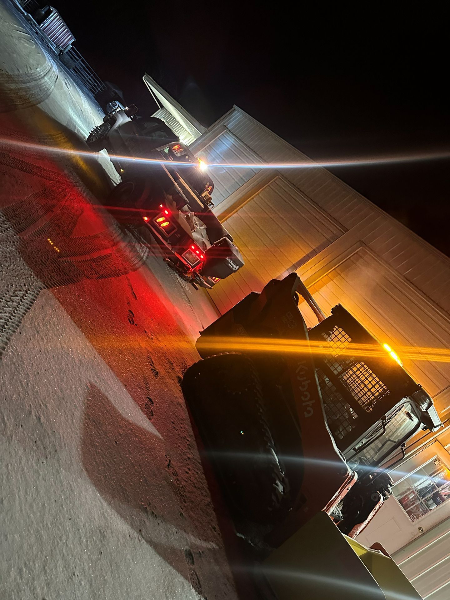 A skid steer loader sits outside a building at night, with a truck parked nearby and bright lights reflecting in the lens.