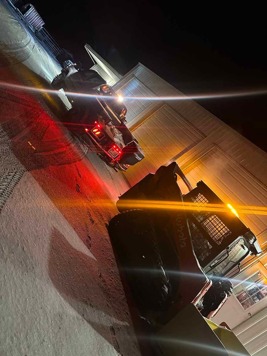 A low-angle night view of a dark tow truck and a car parked on an asphalt lot beside a building under bright streetlights.
