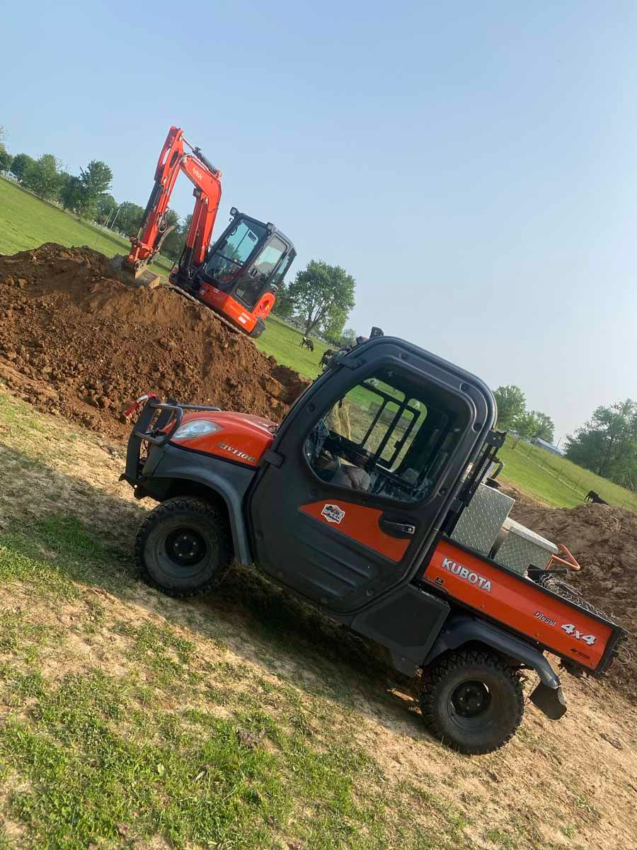 A Kubota utility vehicle parked on a dirt site near a large pile of excavated soil and an orange excavator.