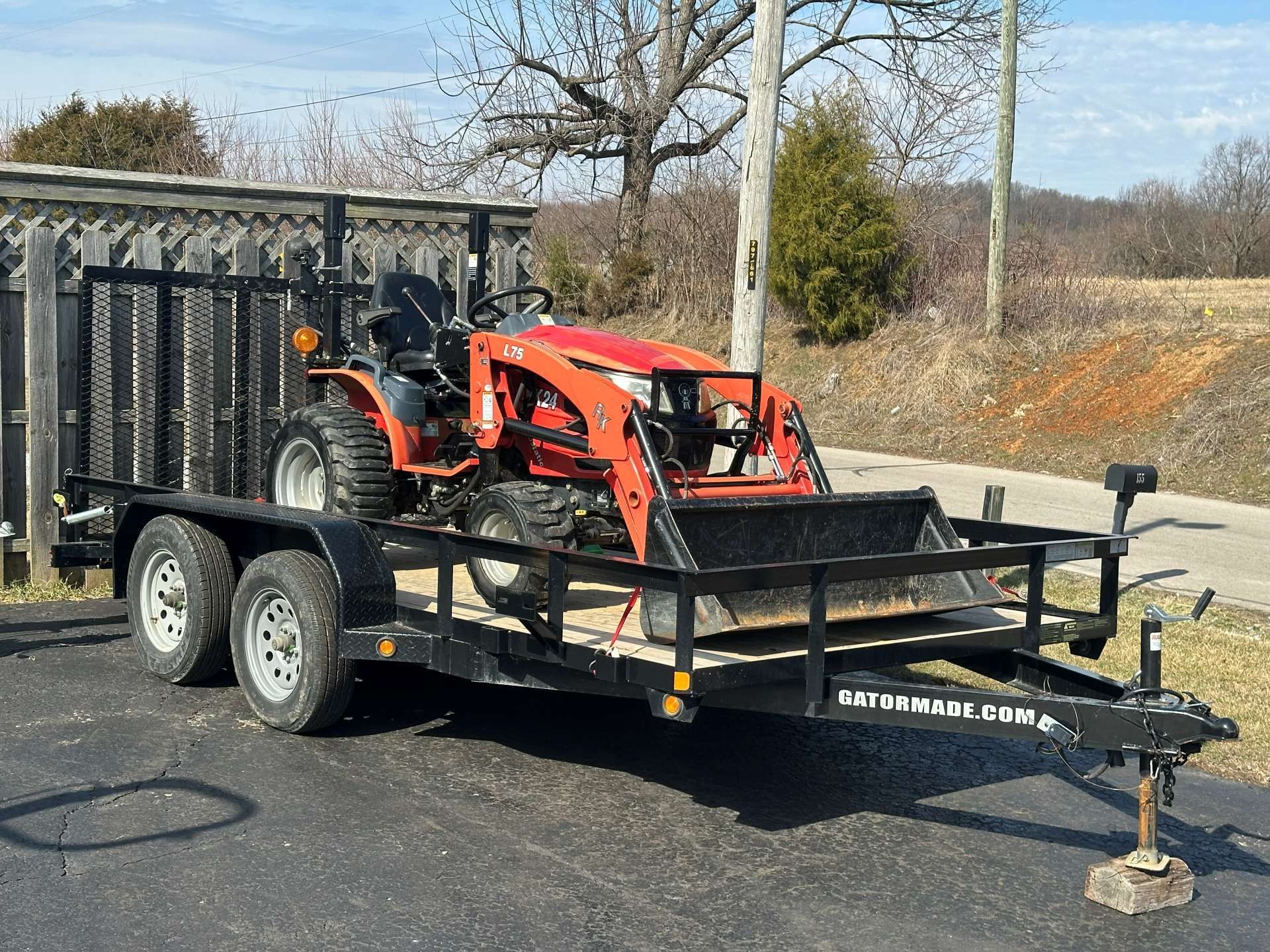 A red Kubota tractor with a front loader is loaded onto a black tandem-axle utility trailer parked on an asphalt surface.