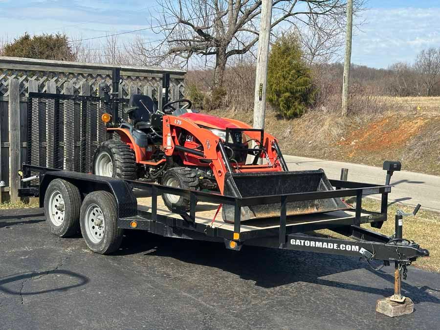 An orange compact tractor with a front loader bucket parked on a flatbed utility trailer on an asphalt surface.