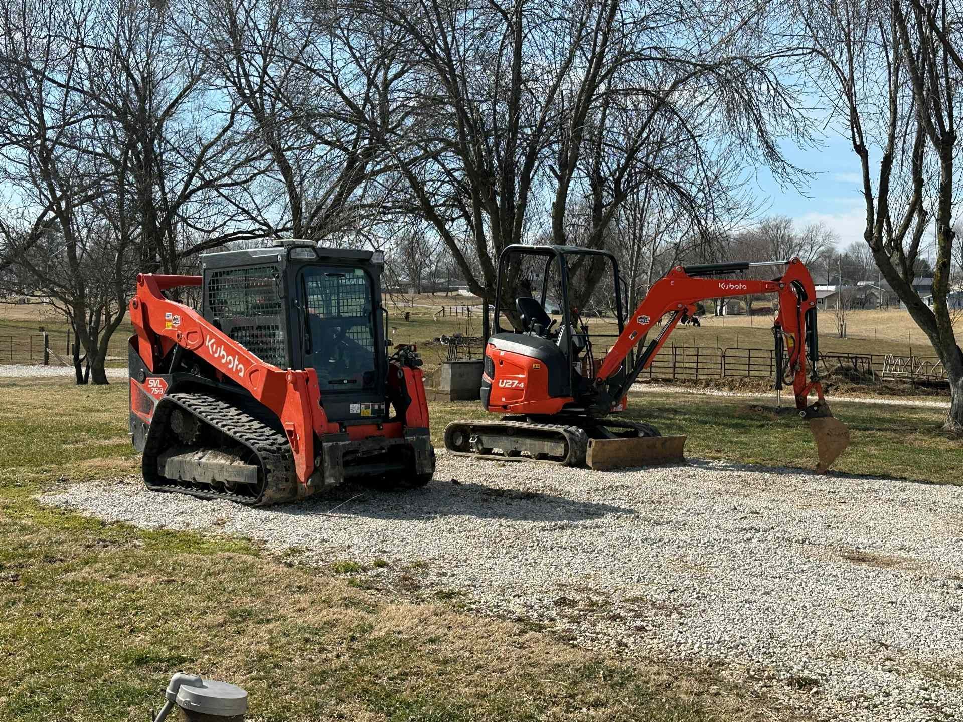 Orange Kubota tracked skid steer and mini excavator parked on a gravel patch in a rural, grassy field under a blue sky.