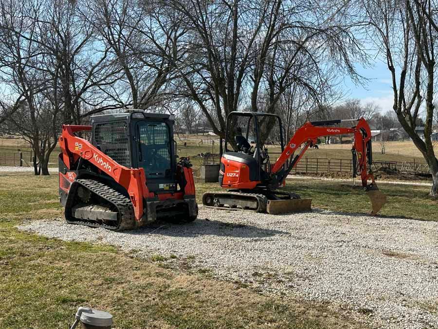 Two bright orange Kubota construction machines, a skid steer and an excavator, parked on a gravel lot under bare trees.