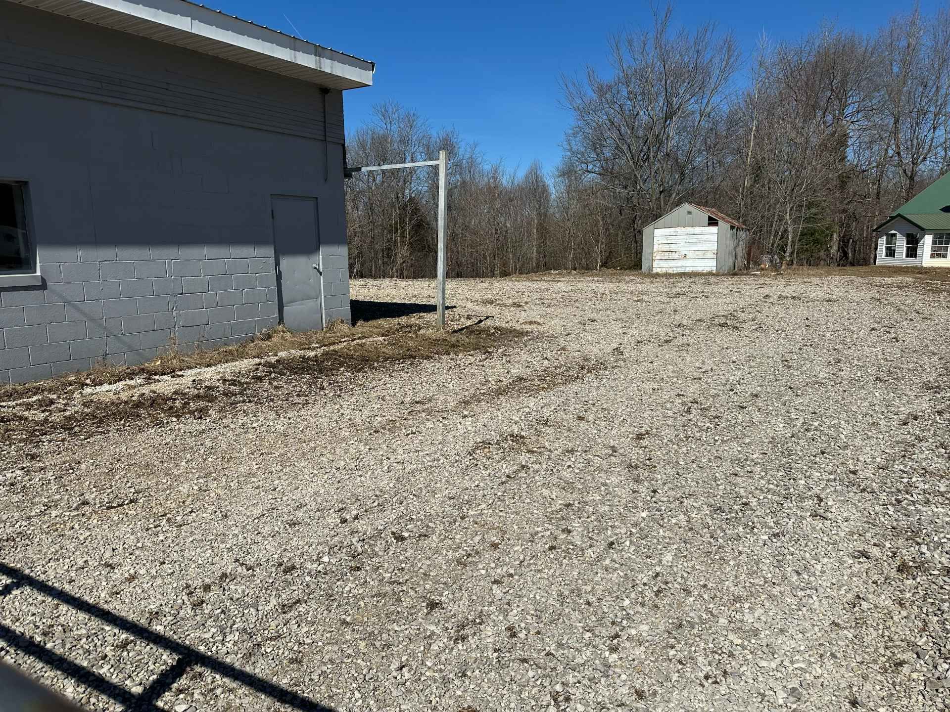 A light-colored building with a gravel lot, a metal post, and distant small structures under a clear blue sky.