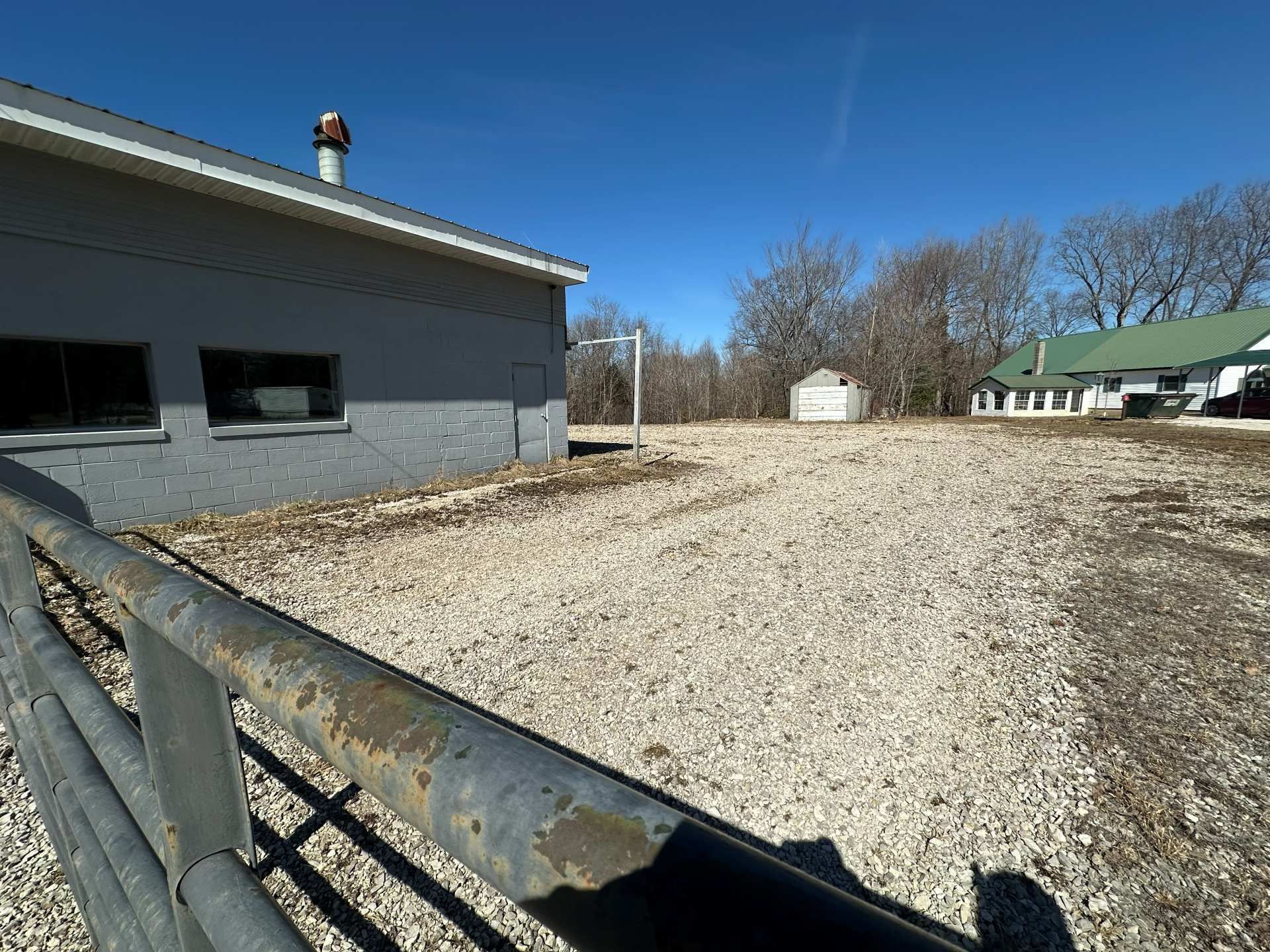 A gravel lot sits between a gray building and a white house, separated by a metal fence in the foreground.