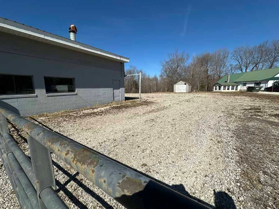 A gravel area between a gray building and a white house, seen from behind a metal gate under a clear blue sky.