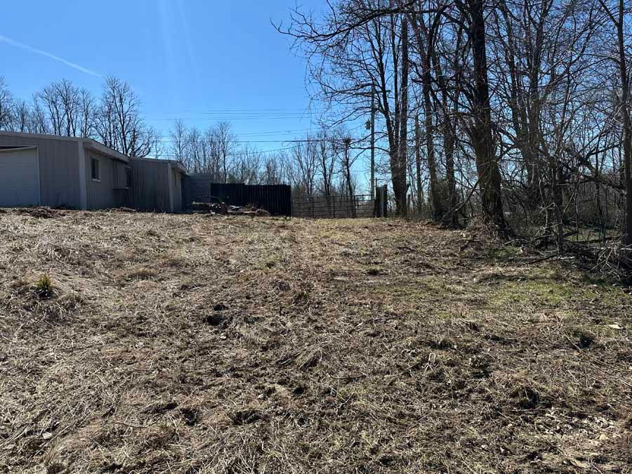 A large, grey outbuilding sits on a clearing with dry grass and dirt, framed by a line of bare trees under a clear sky.