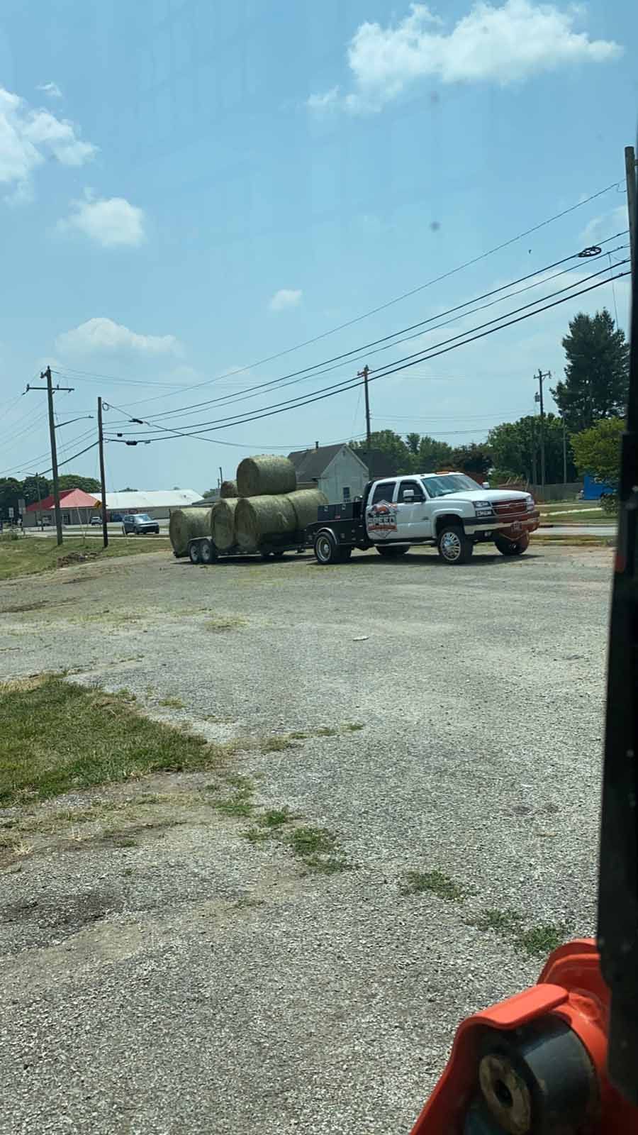 A white pickup truck towing a trailer loaded with several large, round hay bales parked on a gravel lot under a sunny sky.