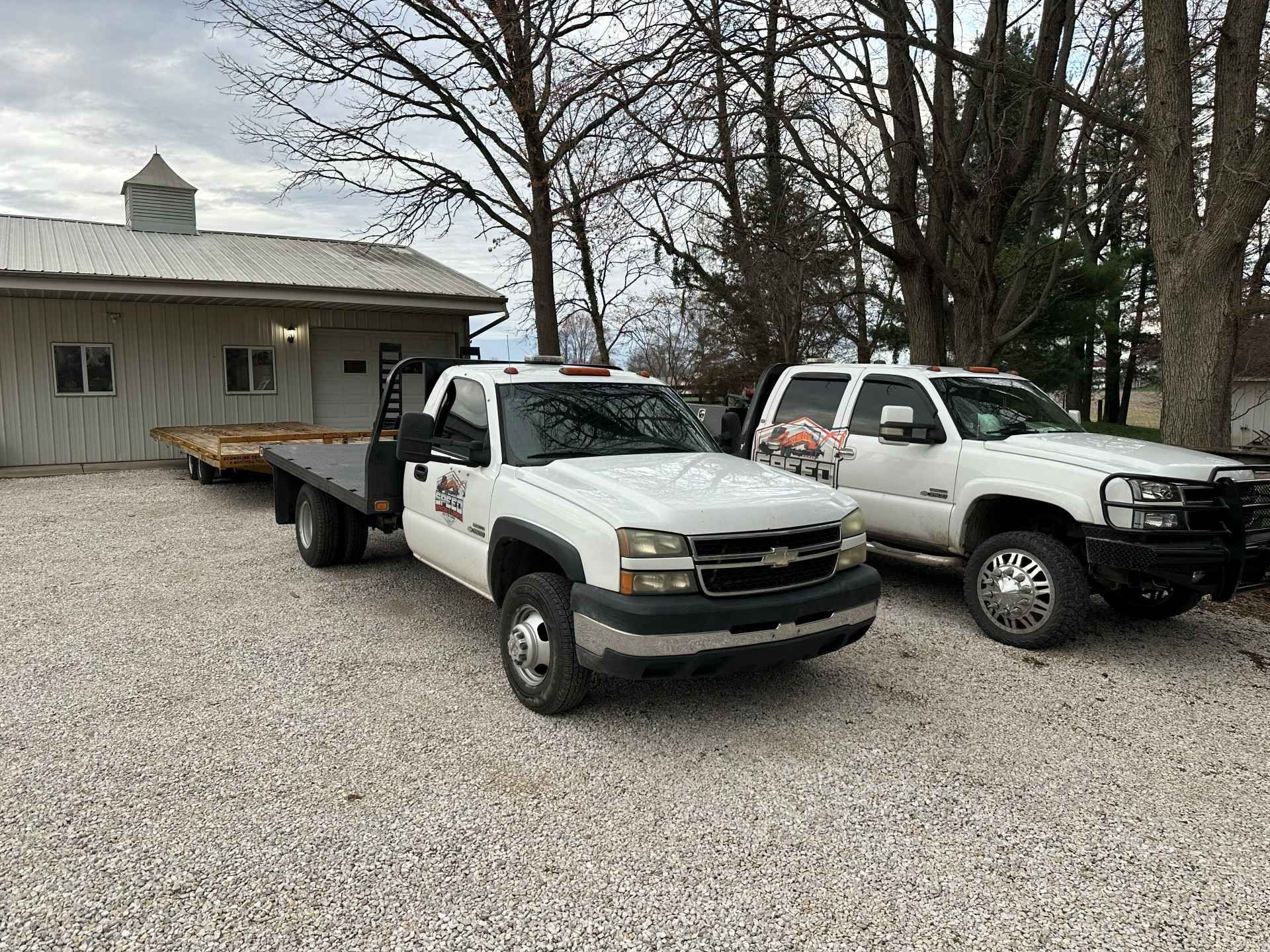 Two white Chevrolet pickup trucks parked on a gravel lot in front of a light-colored barn building.