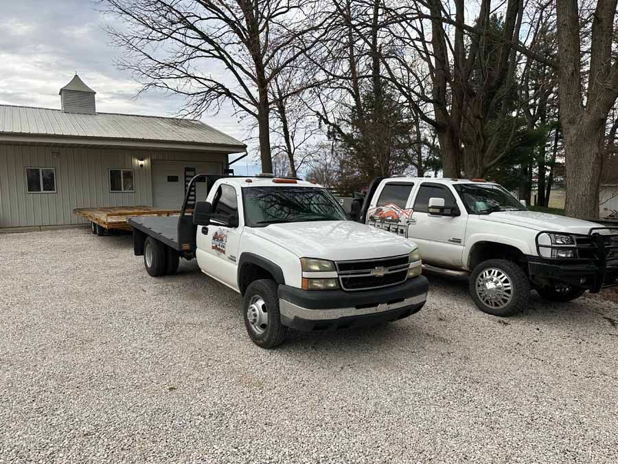 Two white pickup trucks, one with a flatbed trailer, parked on a gravel lot in front of a building.