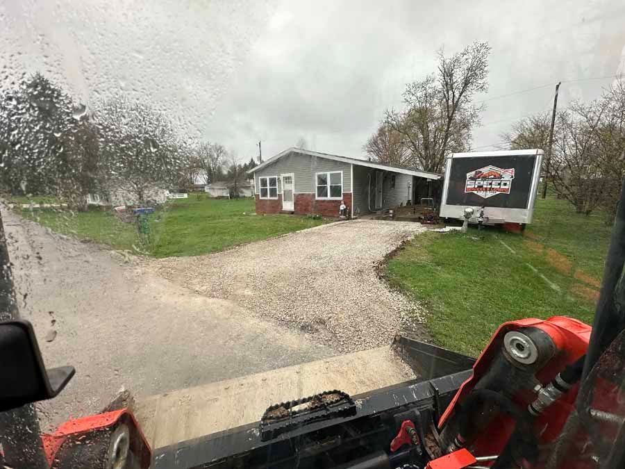 A view from inside heavy equipment showing a gravel driveway leading to a gray and brick house with an attached trailer.