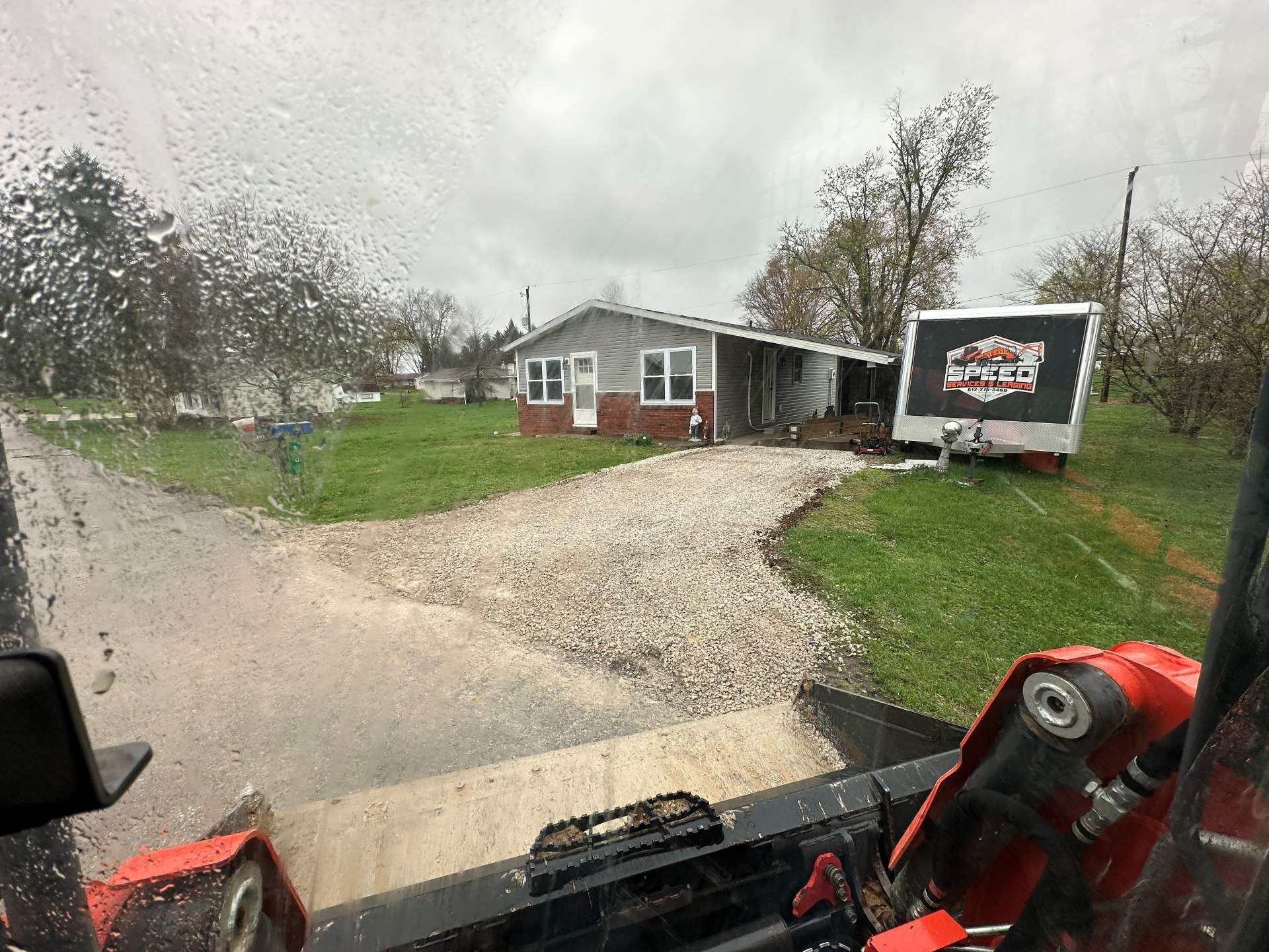 View from a skid steer cab overlooking a gravel driveway leading to a single-story home and a parked trailer.