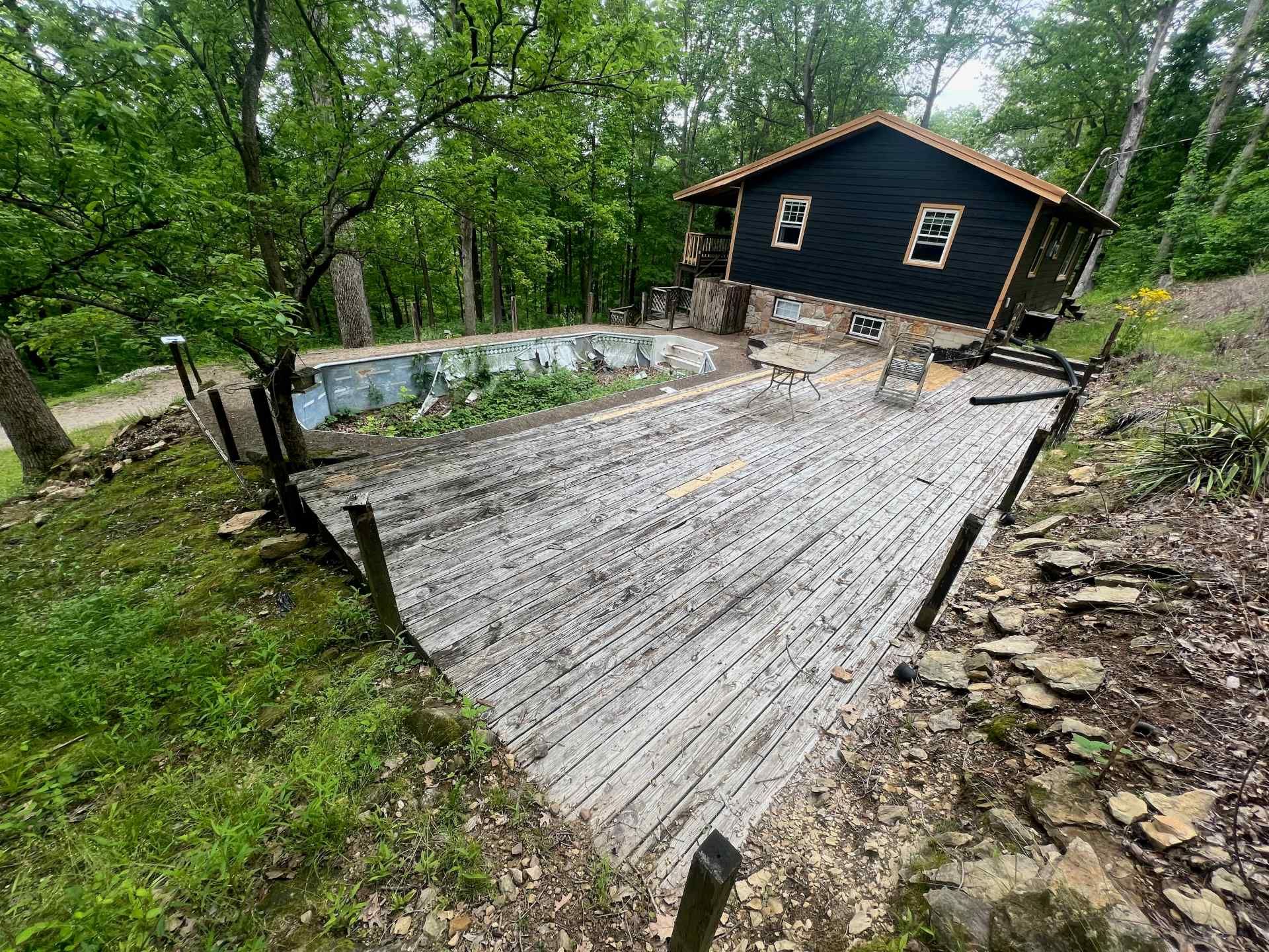 A weathered wooden deck leads to a small, dark-sided cabin nestled in a lush, green, wooded hillside.