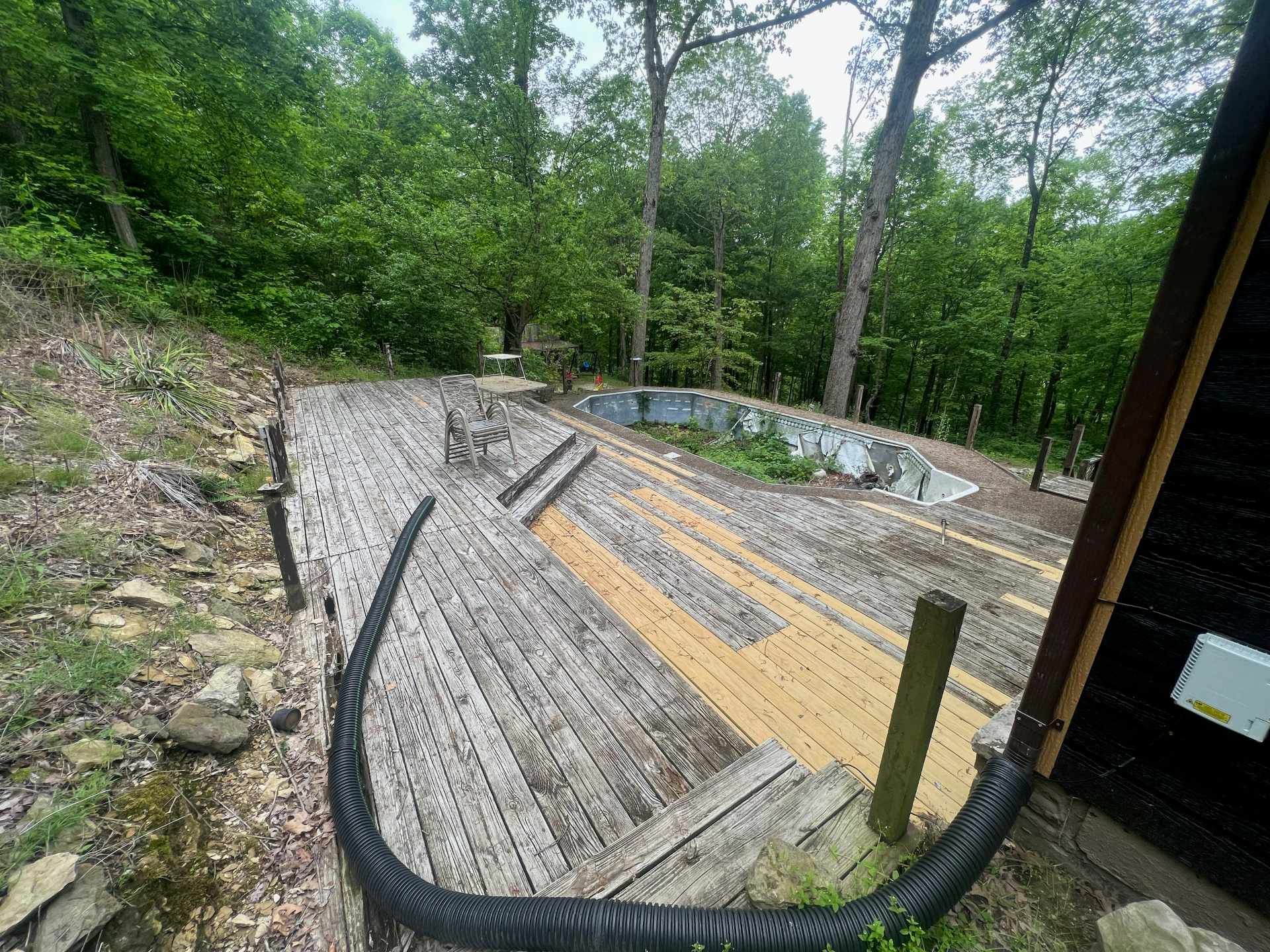 An outdoor wooden deck under construction with a surrounding forest, partially installed boards, and a drainage pipe.