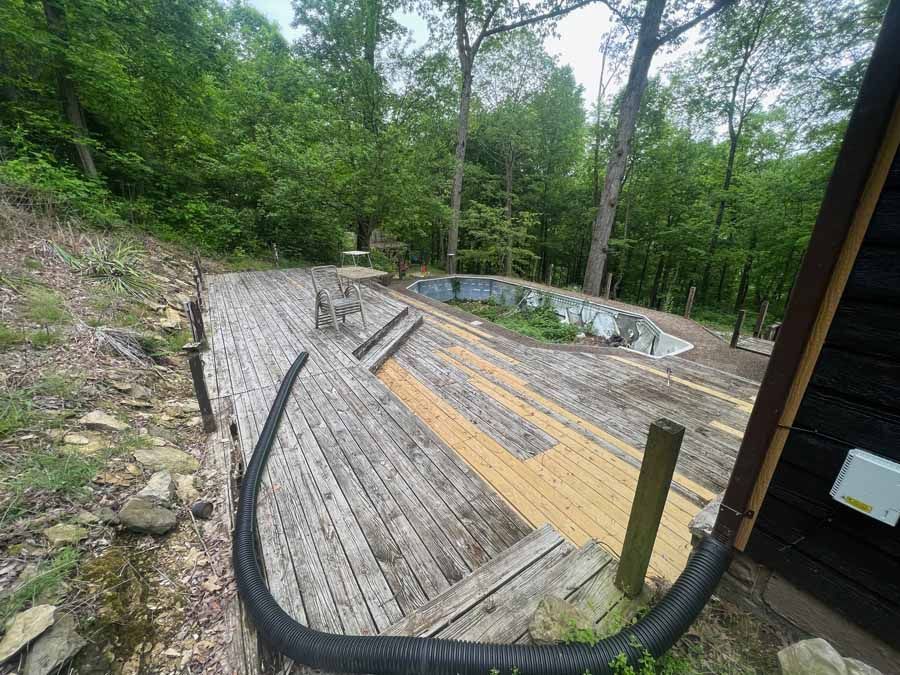 A wooden deck with a mix of weathered gray and new light-colored boards, leading to an overgrown, neglected swimming pool.
