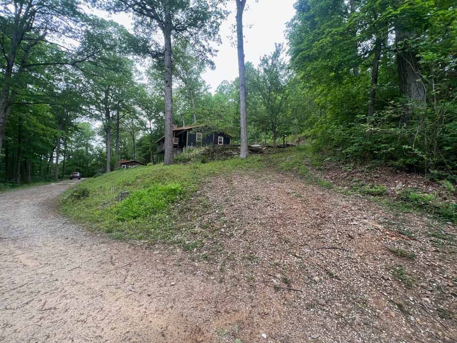 A dirt driveway leads up a grassy slope toward a rustic cabin nestled in a lush, green forest.