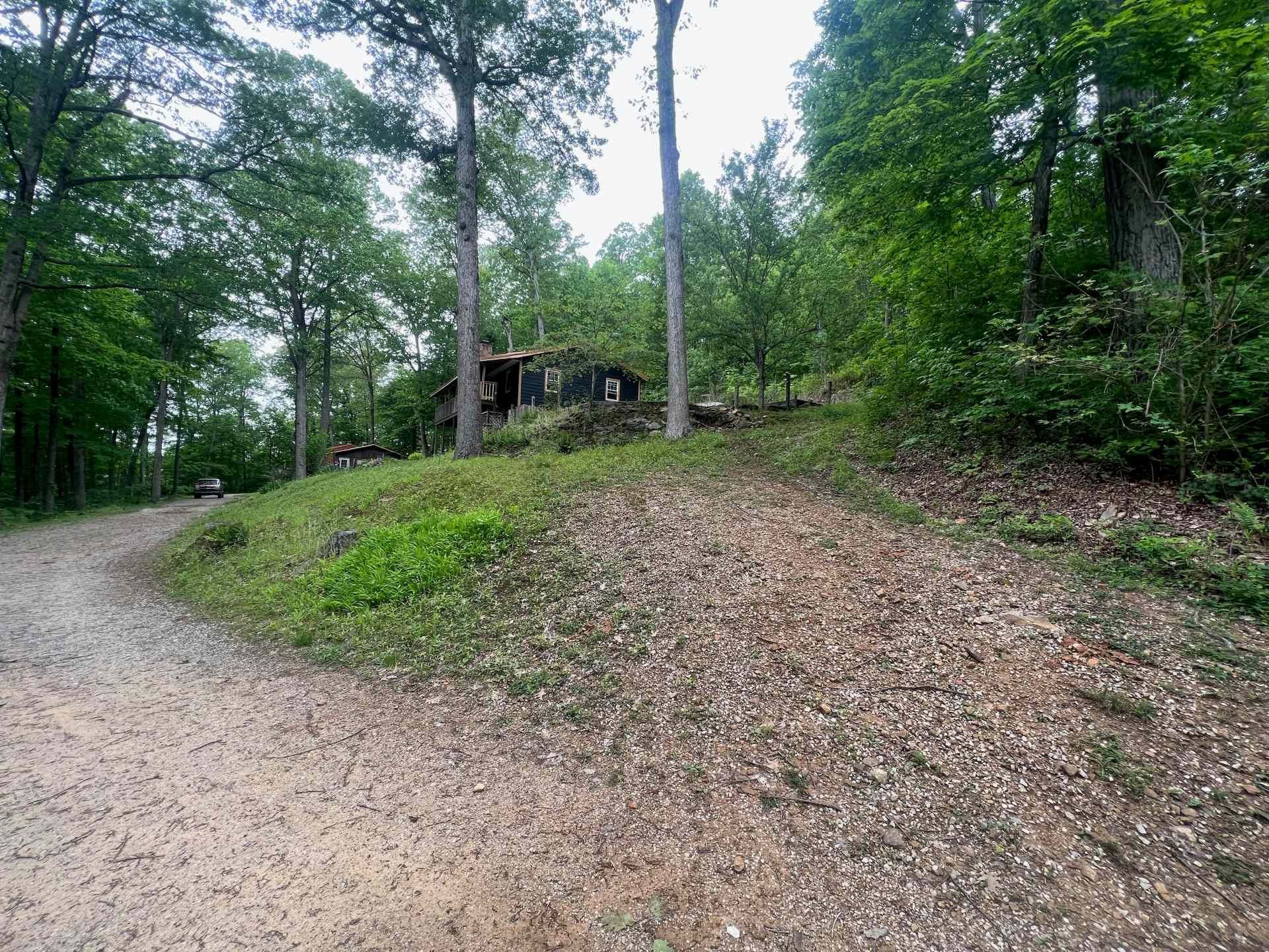 A dark wooden cabin nestled on a wooded hill, overlooking a gravel path winding through a lush, green forest.
