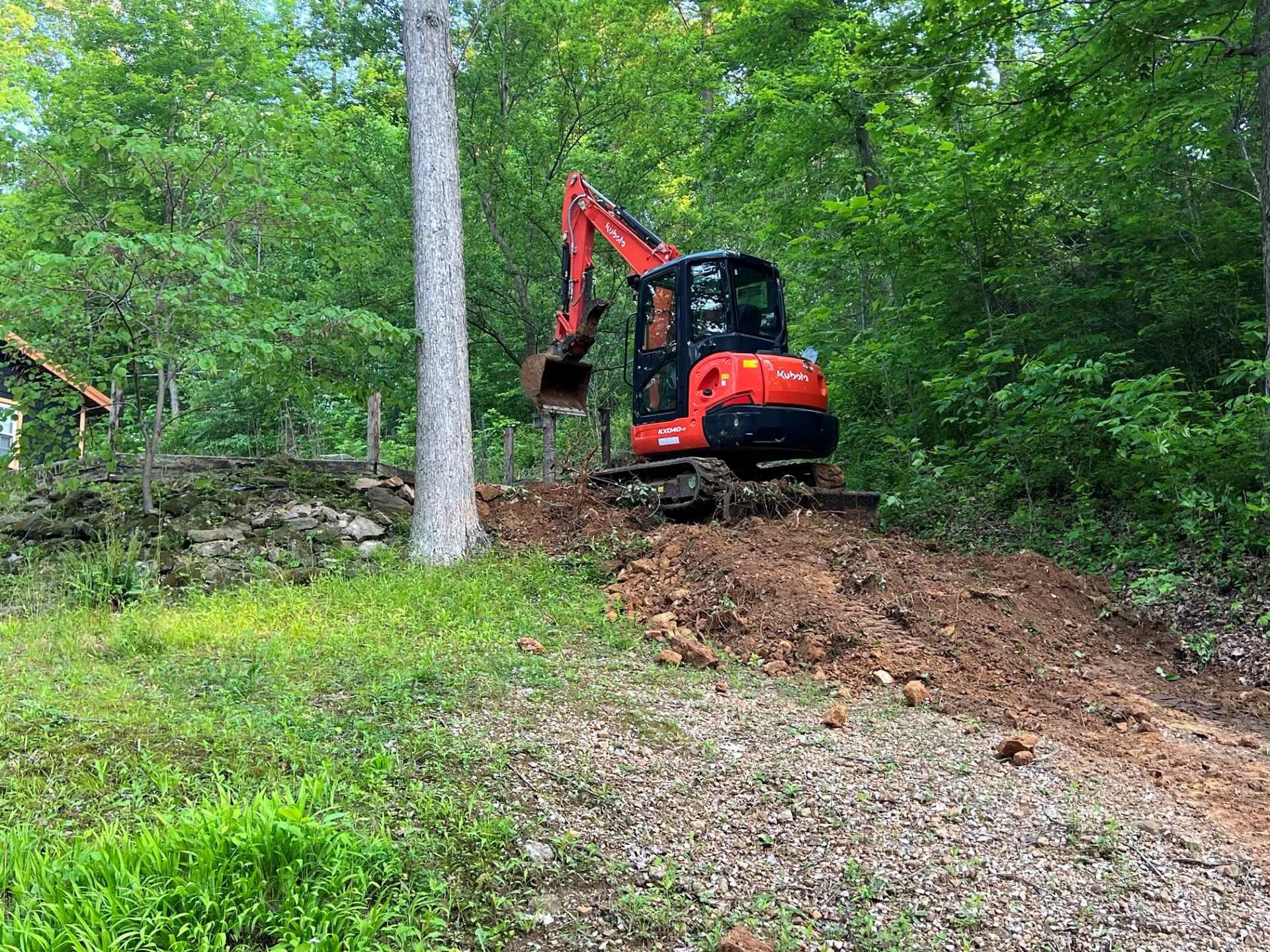 An orange compact excavator parked on a dirt mound in a wooded area.