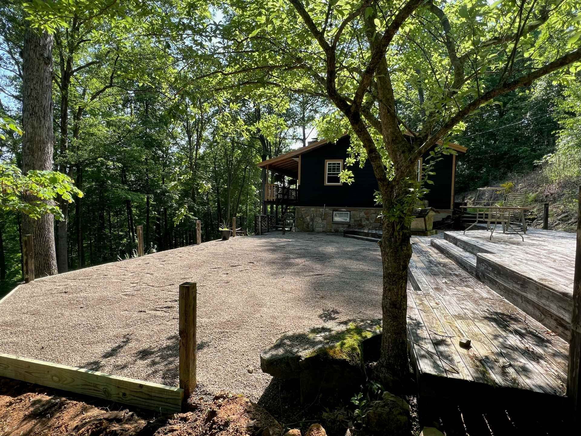 A dark wood cabin sits in a lush forest behind a large, leveled gravel patio area with wooden retaining borders.