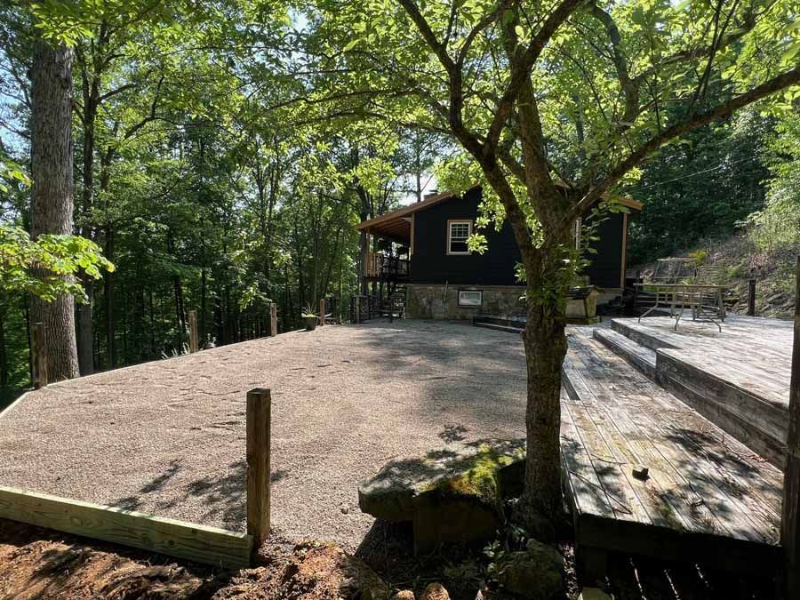 A dark cabin with a stone foundation sits in a forested area next to a gravel patio and a wooden deck.