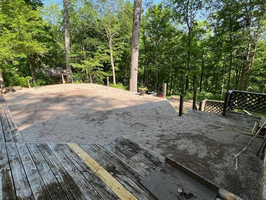 A view from a weathered wooden deck looking out over a newly cleared, light-colored gravel area surrounded by lush trees.