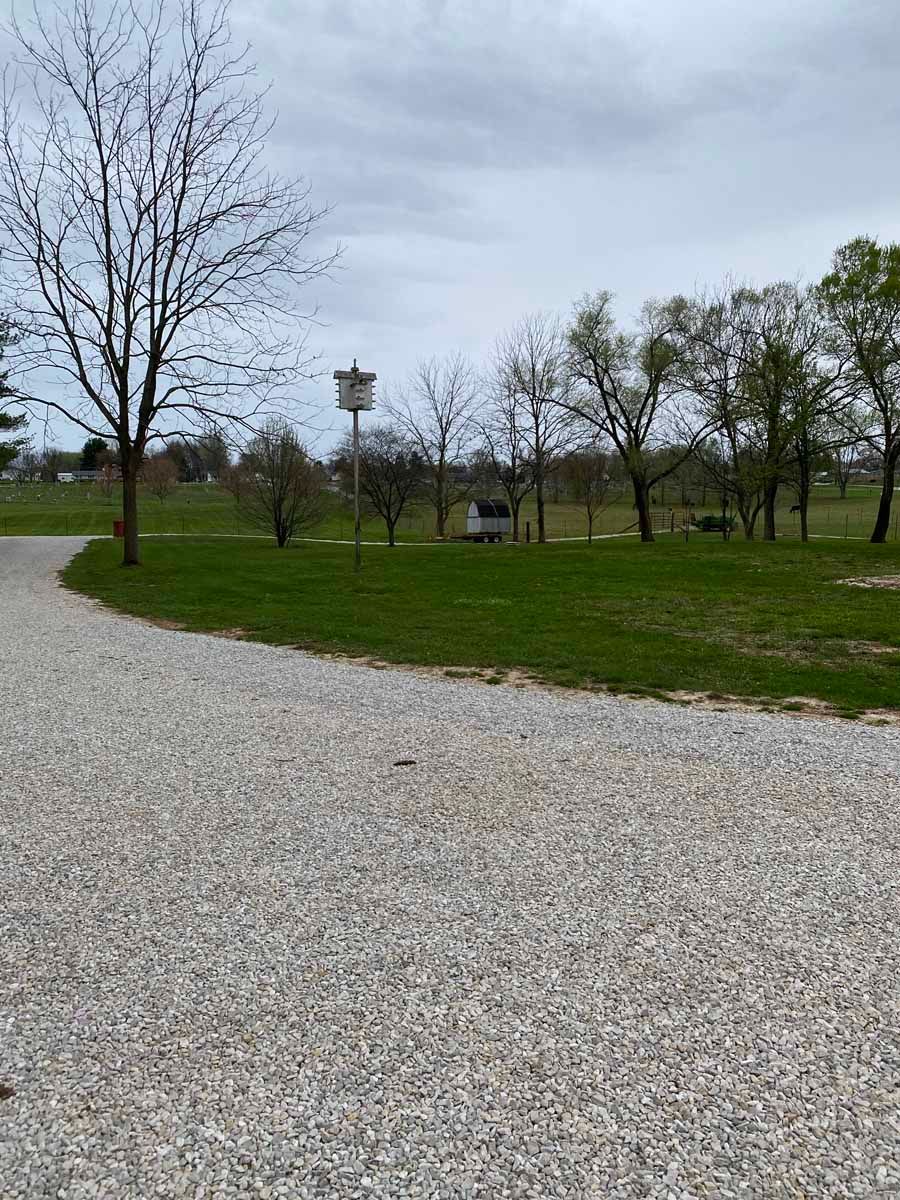 A gravel driveway curves past a grassy lawn featuring a birdhouse on a post and trees under a cloudy sky.