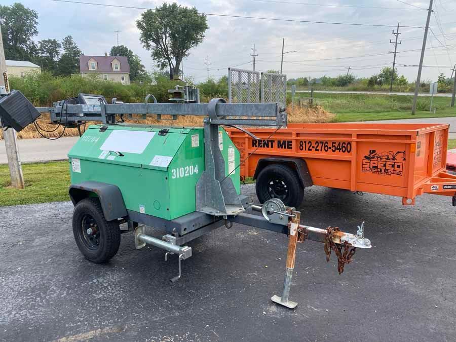 A green portable light tower on a trailer hitch sits next to an orange dump trailer on an asphalt lot.
