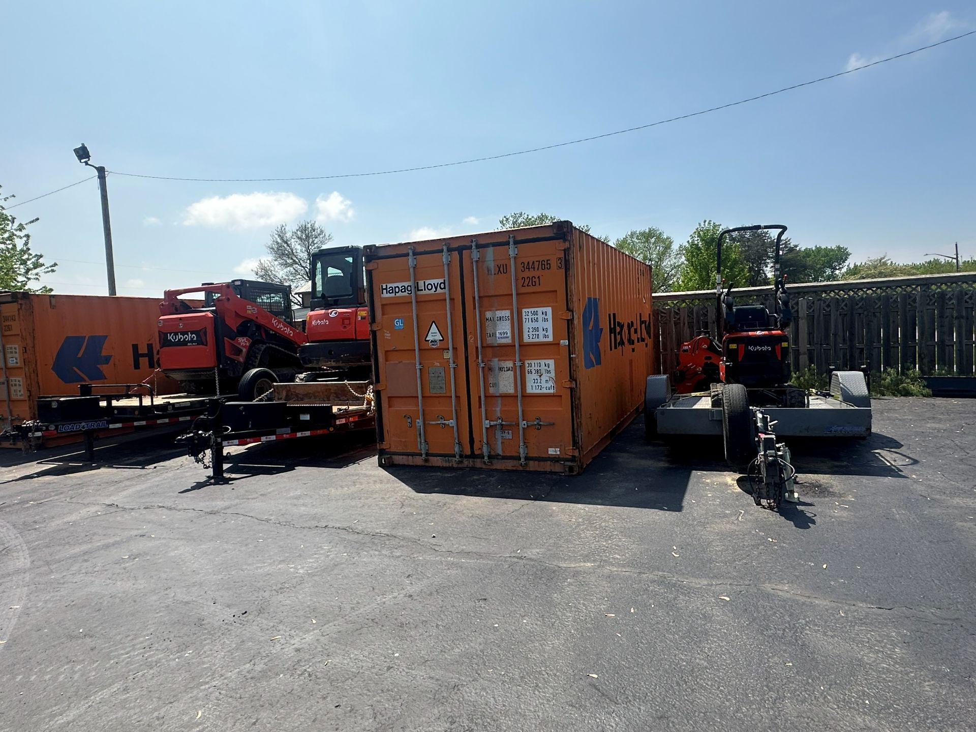 Orange construction containers and pieces of heavy machinery parked on an outdoor asphalt lot under a sunny blue sky.