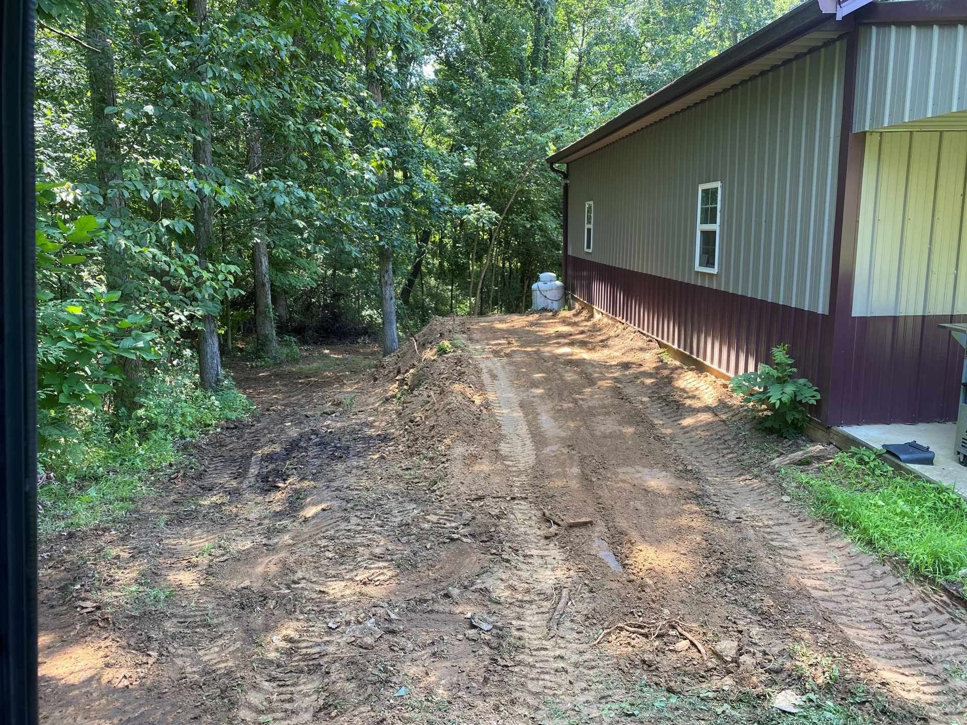 Freshly cleared dirt area beside a building with vertical metal siding, bordered by a dense wooded area.