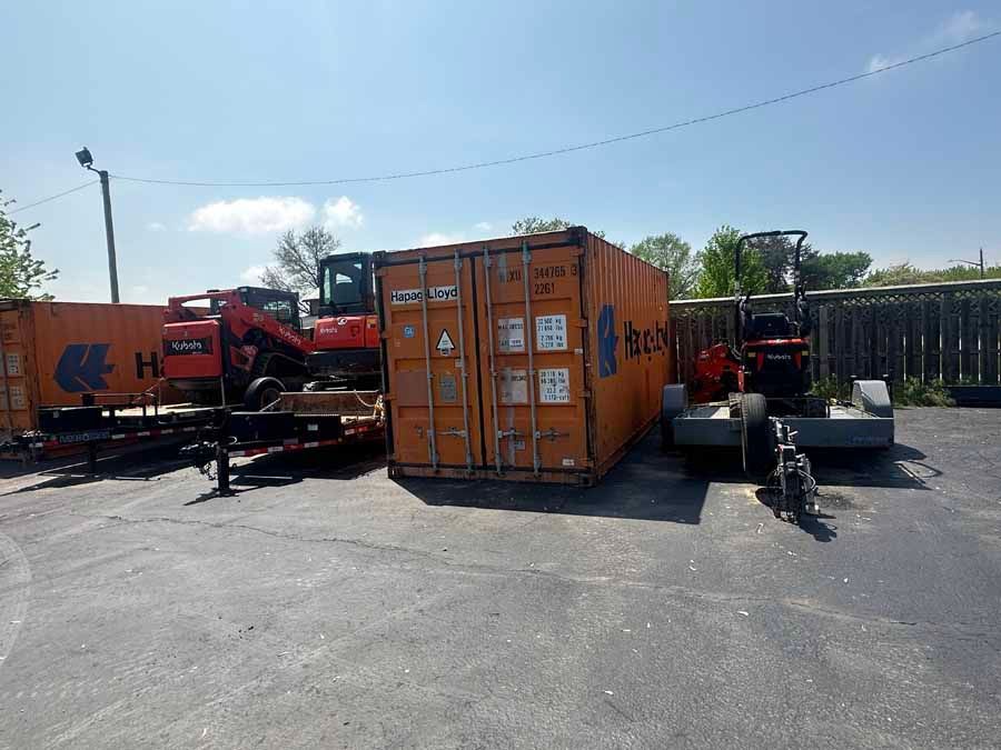 Heavy construction machinery and shipping containers parked on a paved lot under a sunny blue sky.
