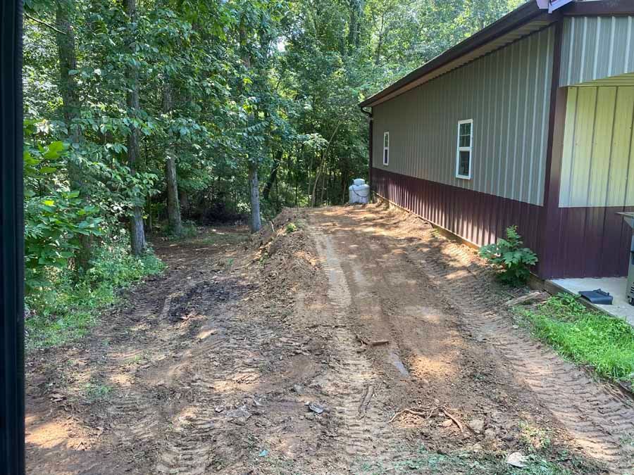 A freshly graded dirt path runs alongside the exterior wall of a metal building, bordering a wooded area.