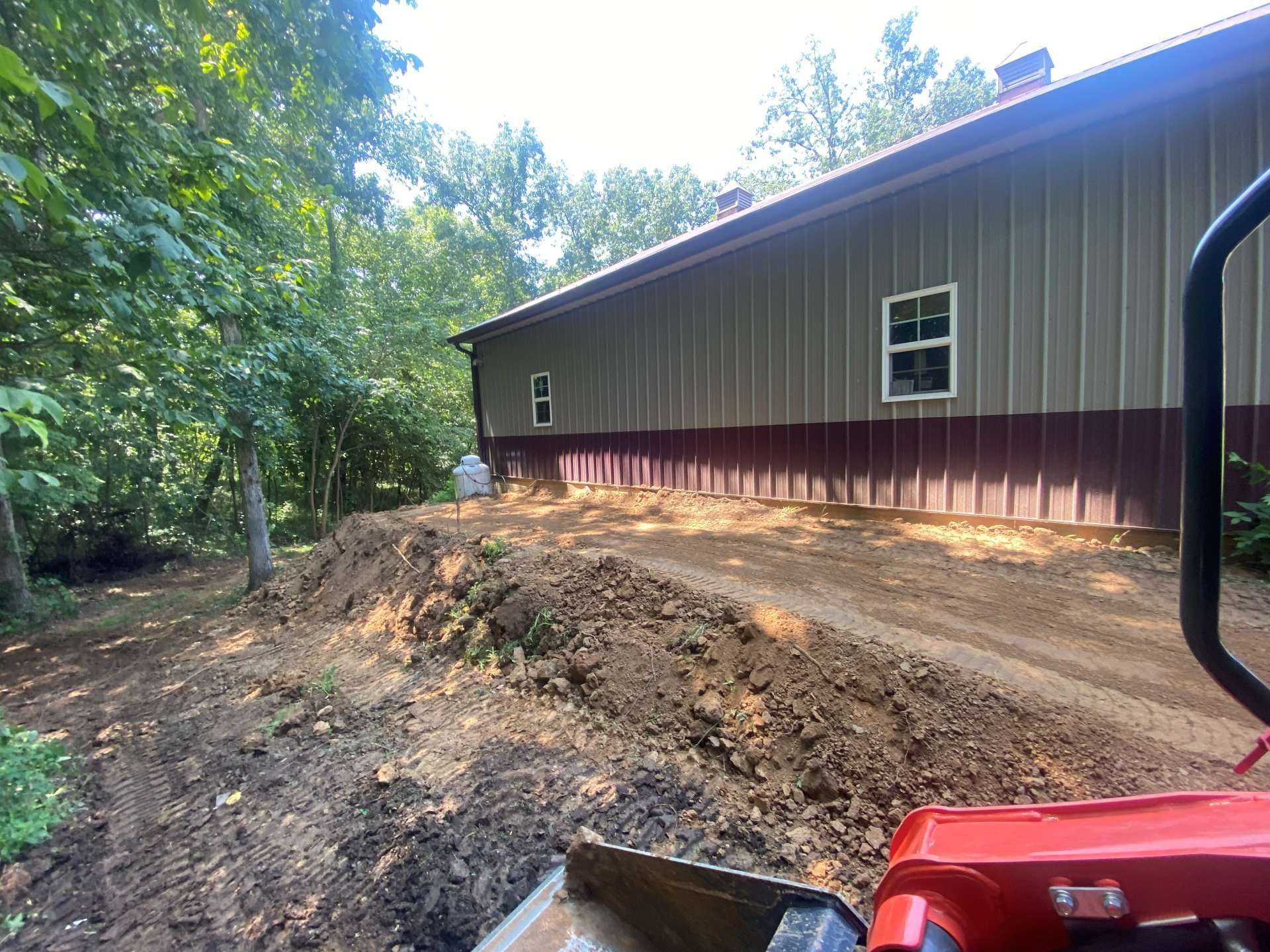 A view from a construction vehicle of a large metal building alongside a freshly excavated dirt slope in a wooded area.