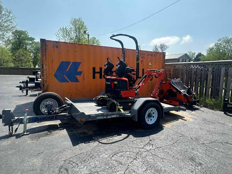 An orange mini-excavator loaded on a flatbed utility trailer parked in a lot next to an orange shipping container.