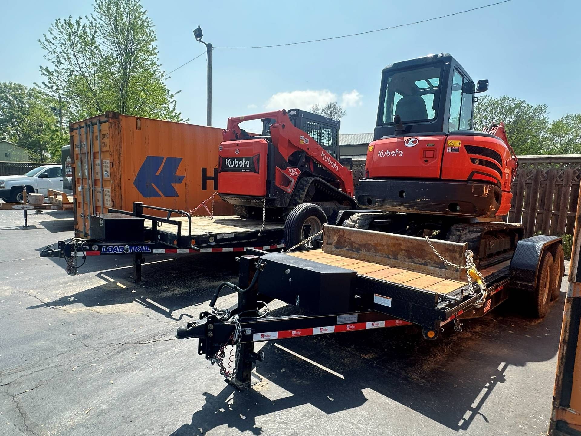 Two orange Kubota construction machines, a skid steer and an excavator, parked on trailers in a sunny outdoor lot.