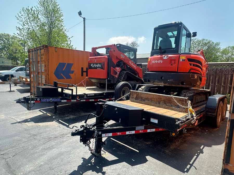 Two orange Kubota construction vehicles on trailers parked outdoors next to a shipping container on a sunny day.