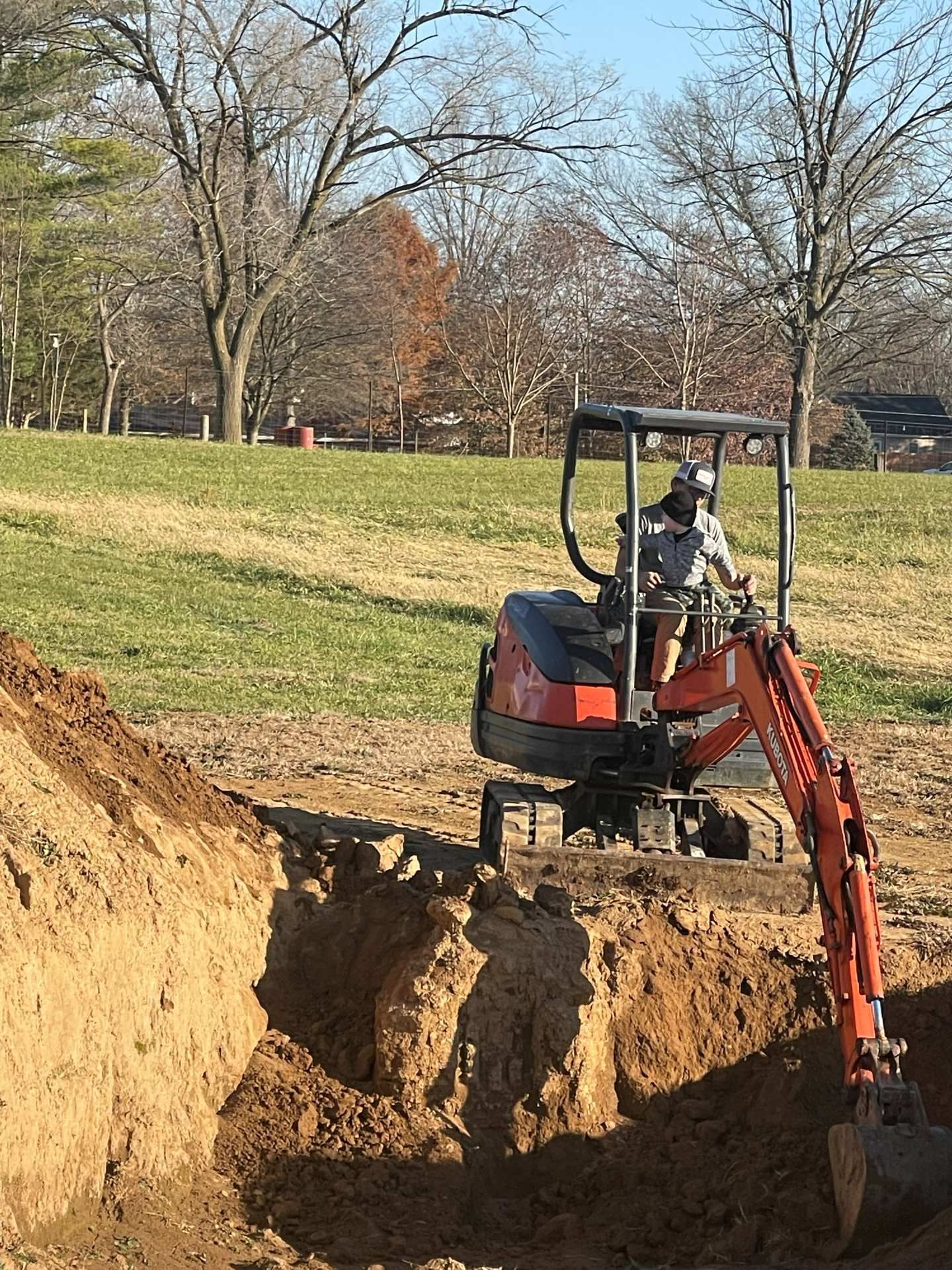 An operator sits in an orange mini excavator, digging a deep trench in an outdoor field.