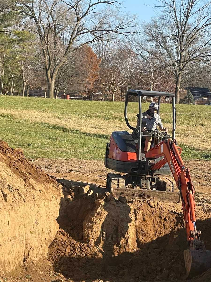 An orange Kubota excavator digs a deep trench in a grassy field under a bright blue sky.