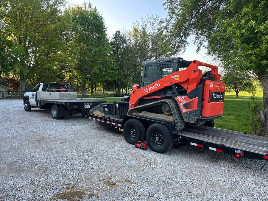 A white flatbed truck hauls a trailer carrying an orange Kubota skid steer loader parked on a gravel lot.